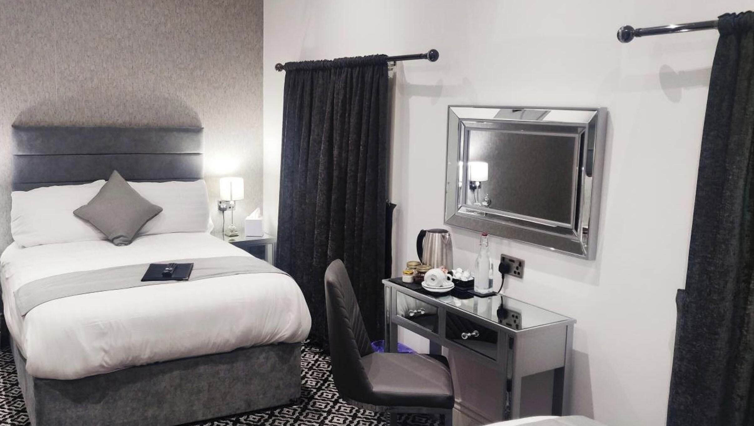 Quarto de hotel com cama, escrivaninha com espelho, cadeira e mesa de cabeceira com abajur.