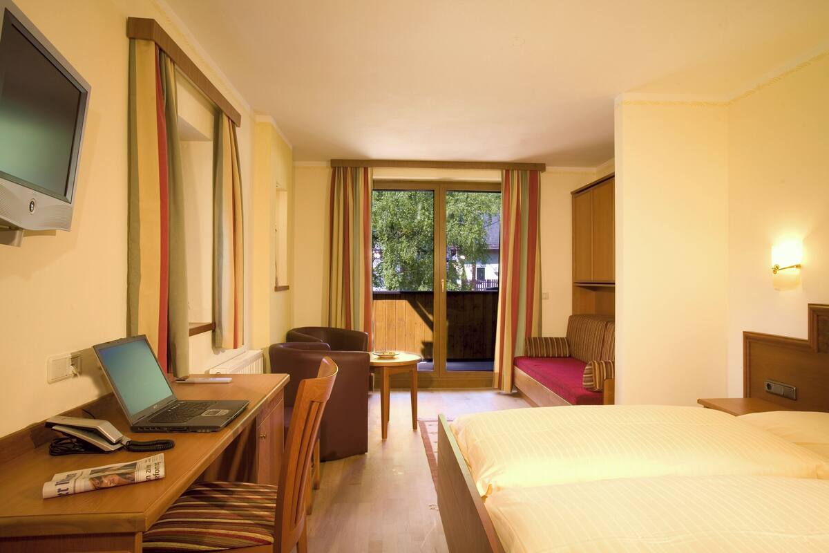 A hotel room with a bed, a desk with a laptop, a chair, a small table, and a TV.