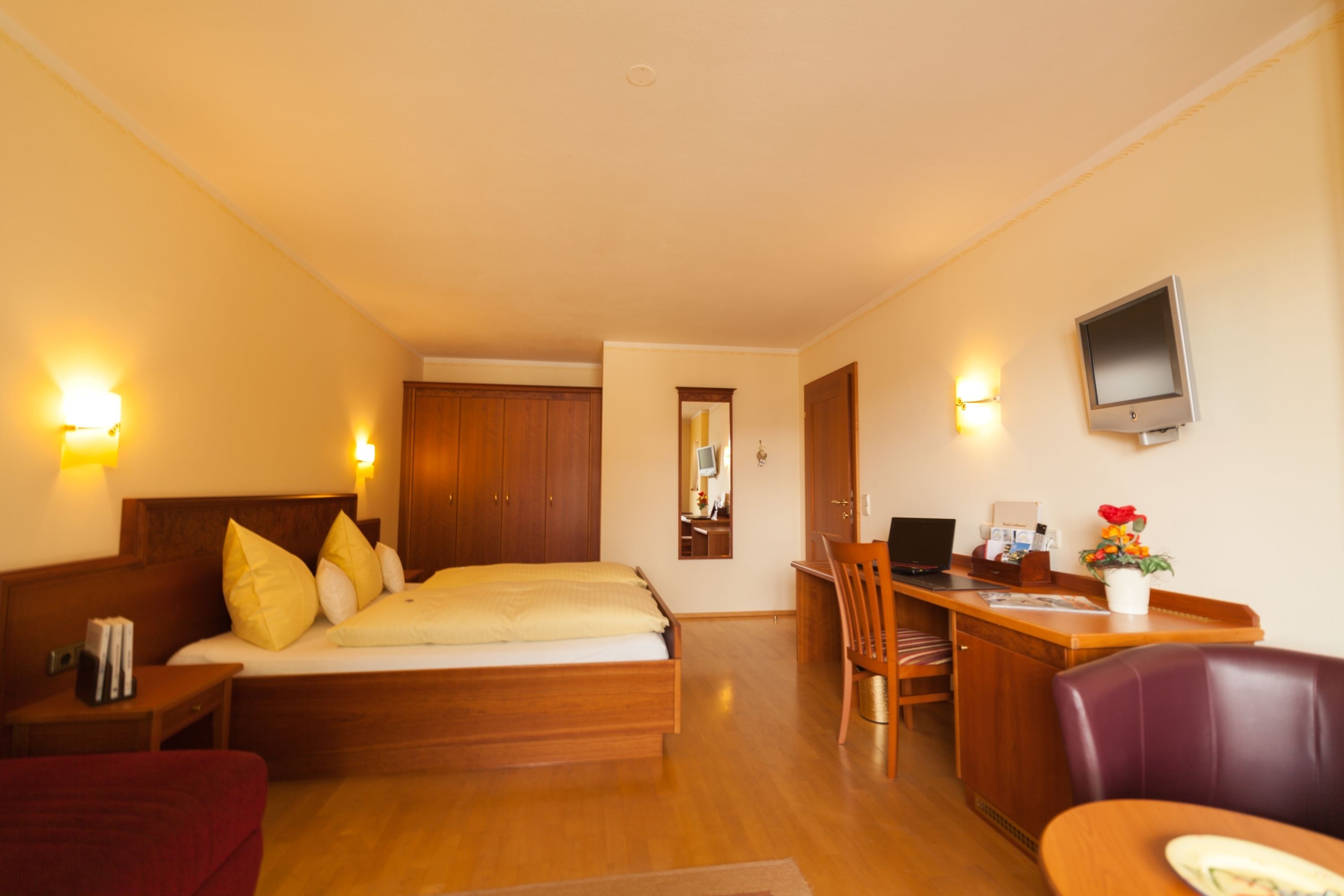 A hotel room with a wooden bed, a desk with a computer, a TV, and a seating area with a chair and a couch.