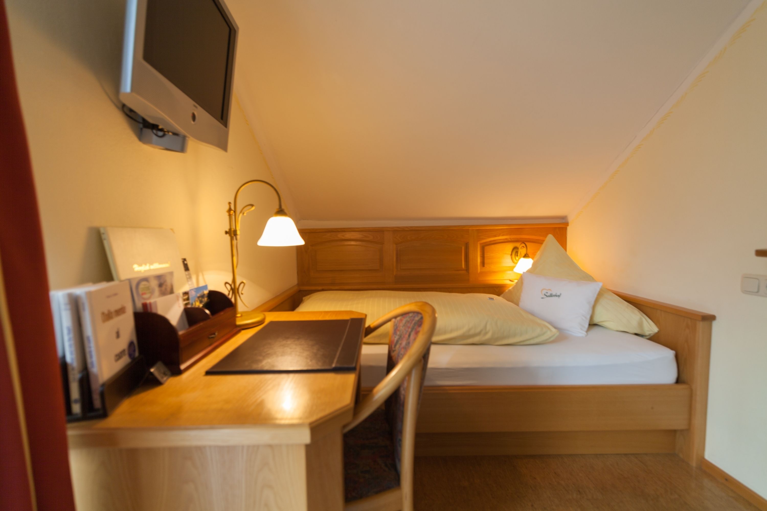 A small, cozy room with a bed, a desk, and a TV mounted on the wall.