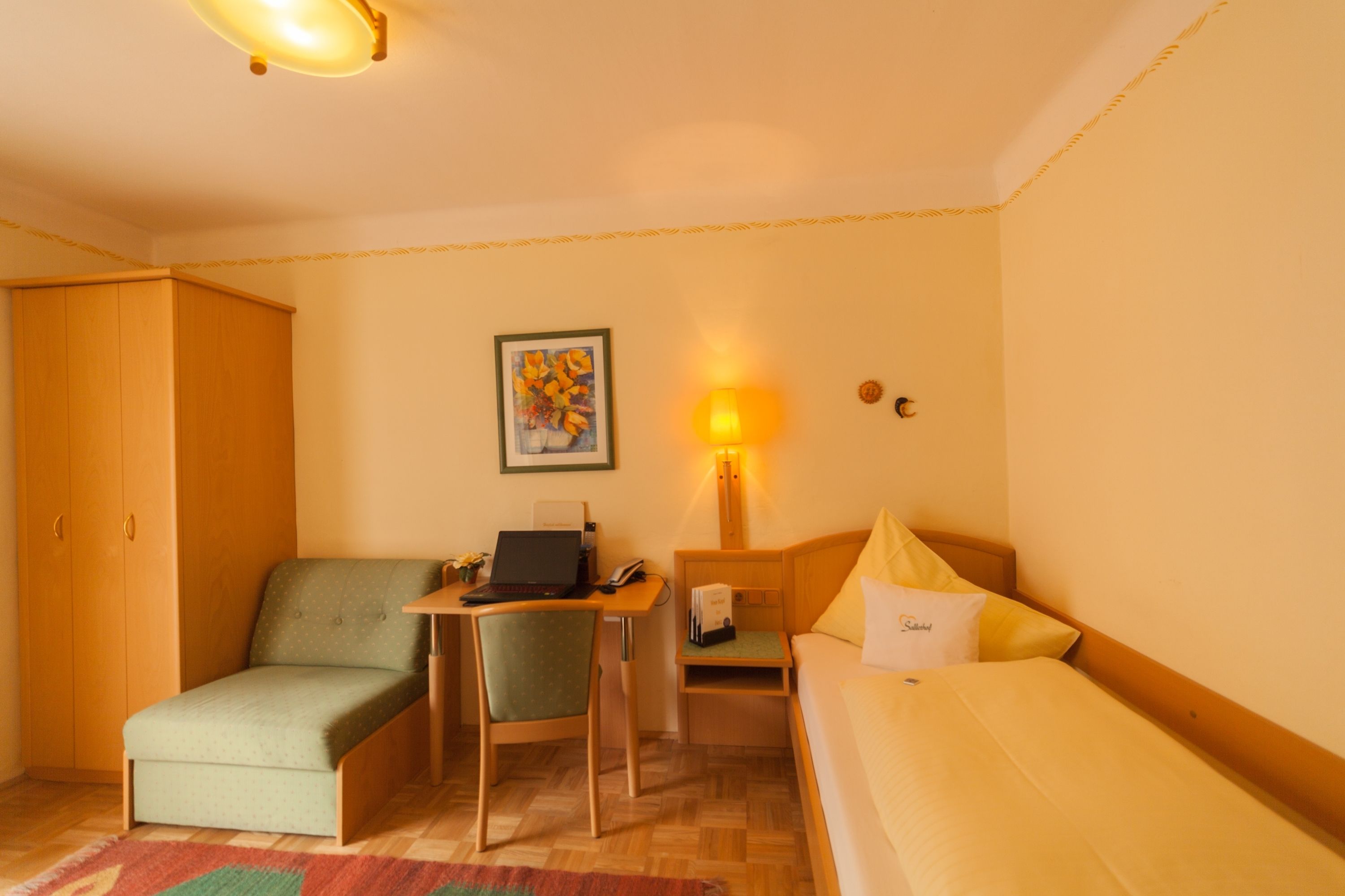 A compact hotel room with a bed, a small desk with a laptop, a chair, a nightstand, and a lamp.
