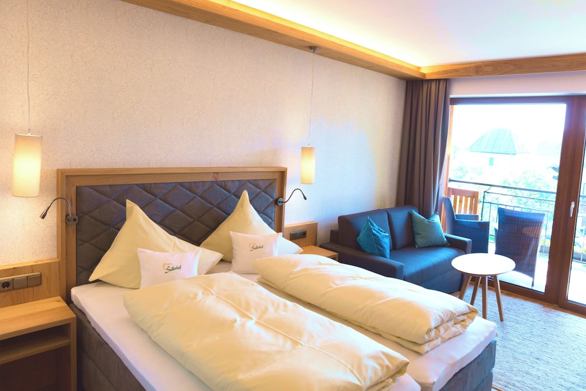 A hotel room with a large bed, a sofa, a small table, and a balcony with outdoor furniture.