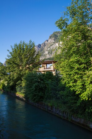 Una cabaña junto al lago con balcón que da a un río, rodeada de abundante vegetación y montañas.
