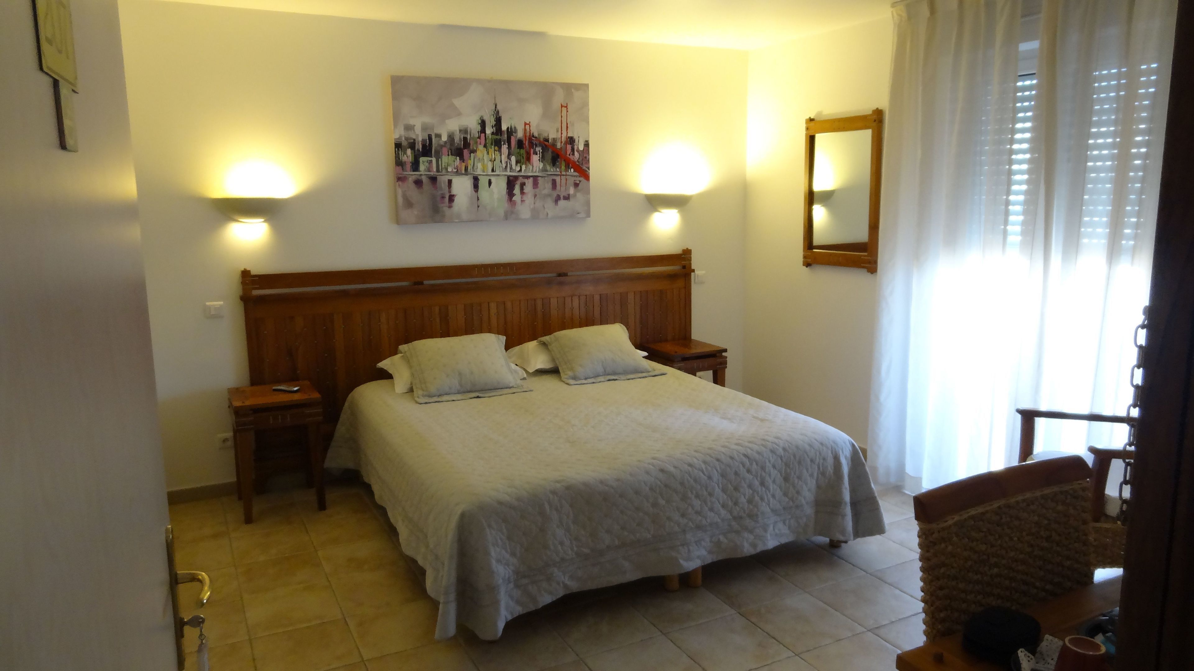 Una habitación de hotel con una cama grande, dos mesitas de noche, un cabecero, un cuadro en la pared y una ventana con cortinas.