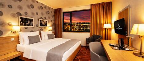 A hotel room with a large bed, a desk with a television, and a city view through the window.