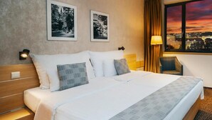 A hotel room with a large bed, two framed pictures on the wall, a chair, and a window with a city view. - Iris Hotel Eden - Czech Leading Hotels (Prague)
