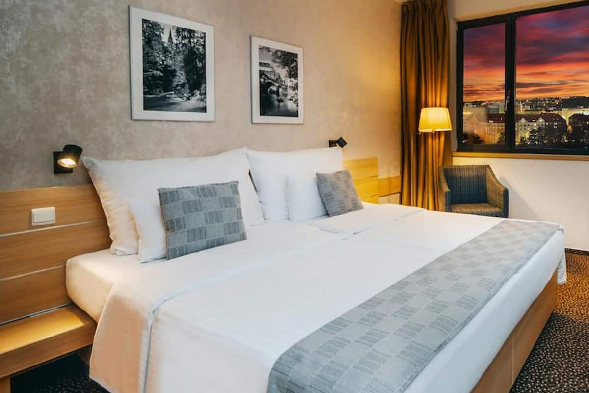 Une chambre d’hôtel avec un grand lit, deux tableaux encadrés au mur, une chaise et une fenêtre donnant sur la ville.