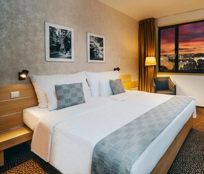 Een hotelkamer met een groot bed, twee ingelijste foto’s aan de muur, een stoel en een raam met uitzicht over de stad.