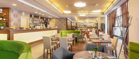 A modern restaurant with round tables, green upholstered chairs, and a bar area with shelves of bottles.