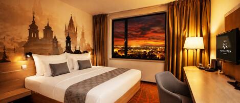 Quarto de hotel com uma cama grande, uma escrivaninha, uma televisão e vista para a paisagem urbana ao pôr do sol.