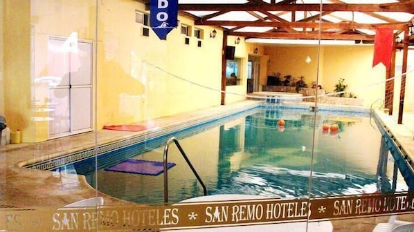 An indoor swimming pool with clear water, surrounded by a glass wall. There are signs indicating 'San Remo Hotel' and a 'DO' sign above the pool.