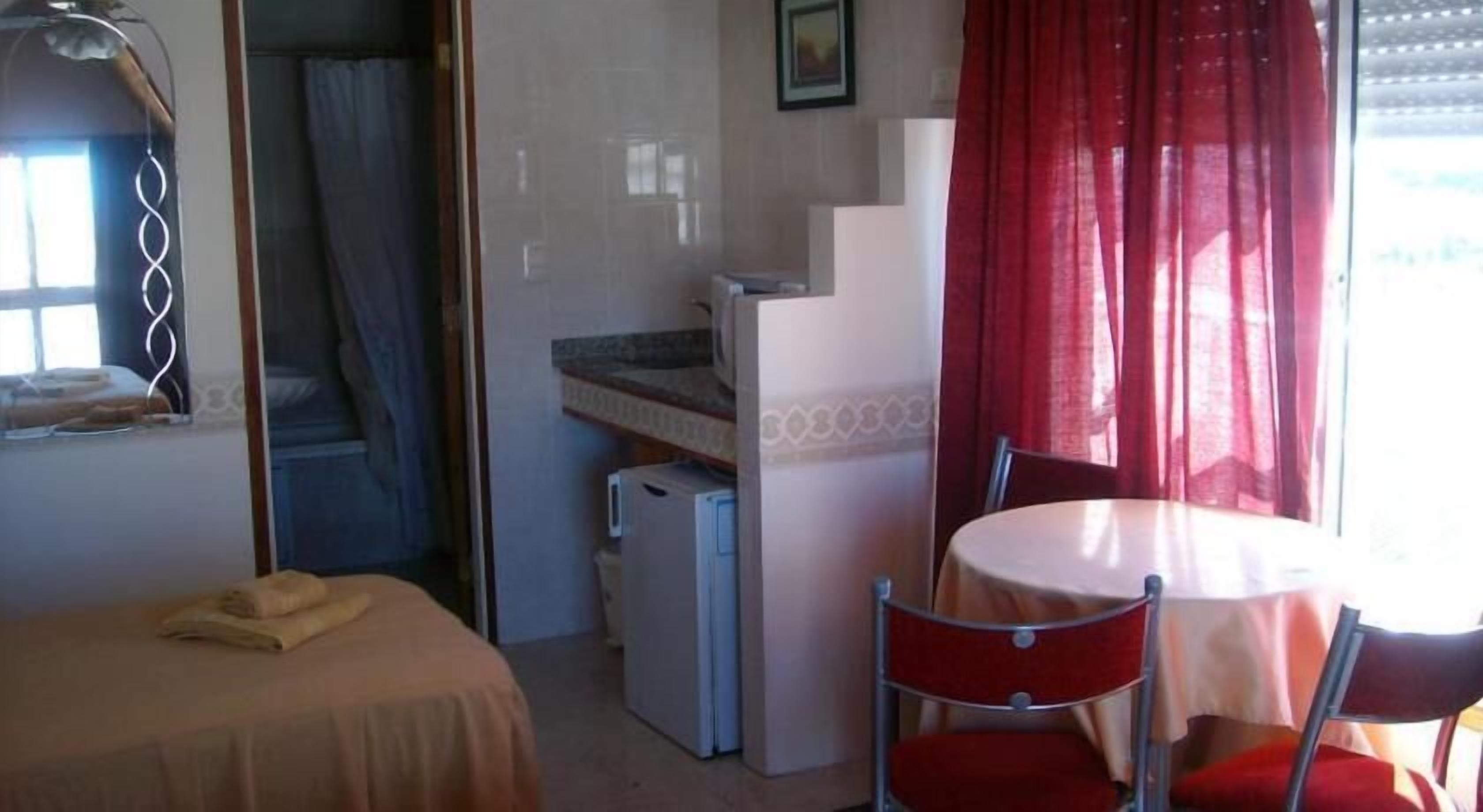 Il s’agit d’une pièce de taille modeste, comprenant un lit, une petite table et une chaise. Un coin cuisine est aménagé avec un réfrigérateur et un four à micro-ondes.