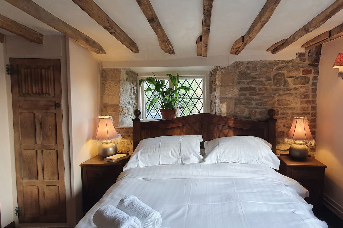 A bedroom with a wooden bed, stone walls, a window with a view, and two bedside lamps.