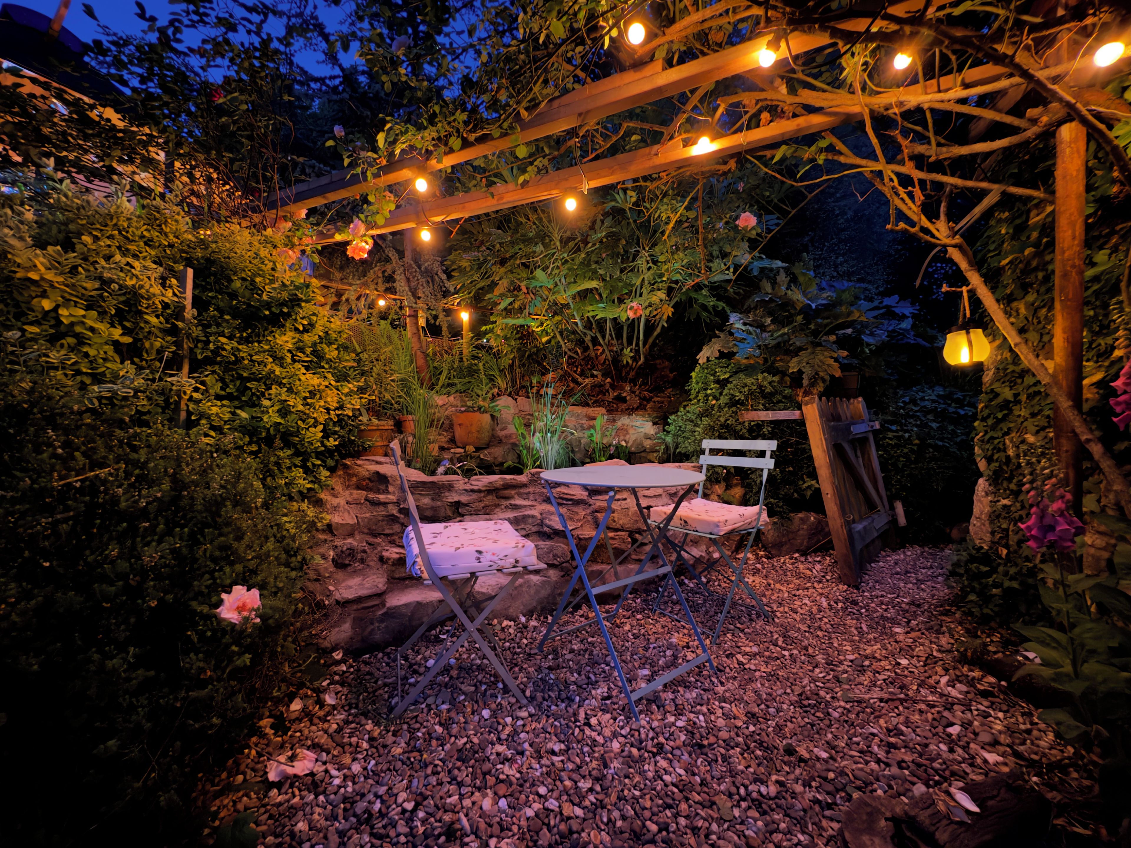 A garden with a wooden pergola, a table, and chairs, surrounded by plants and rocks.