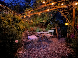 Un jardín con un porche de madera, una mesa y sillas, rodeado de plantas y rocas.