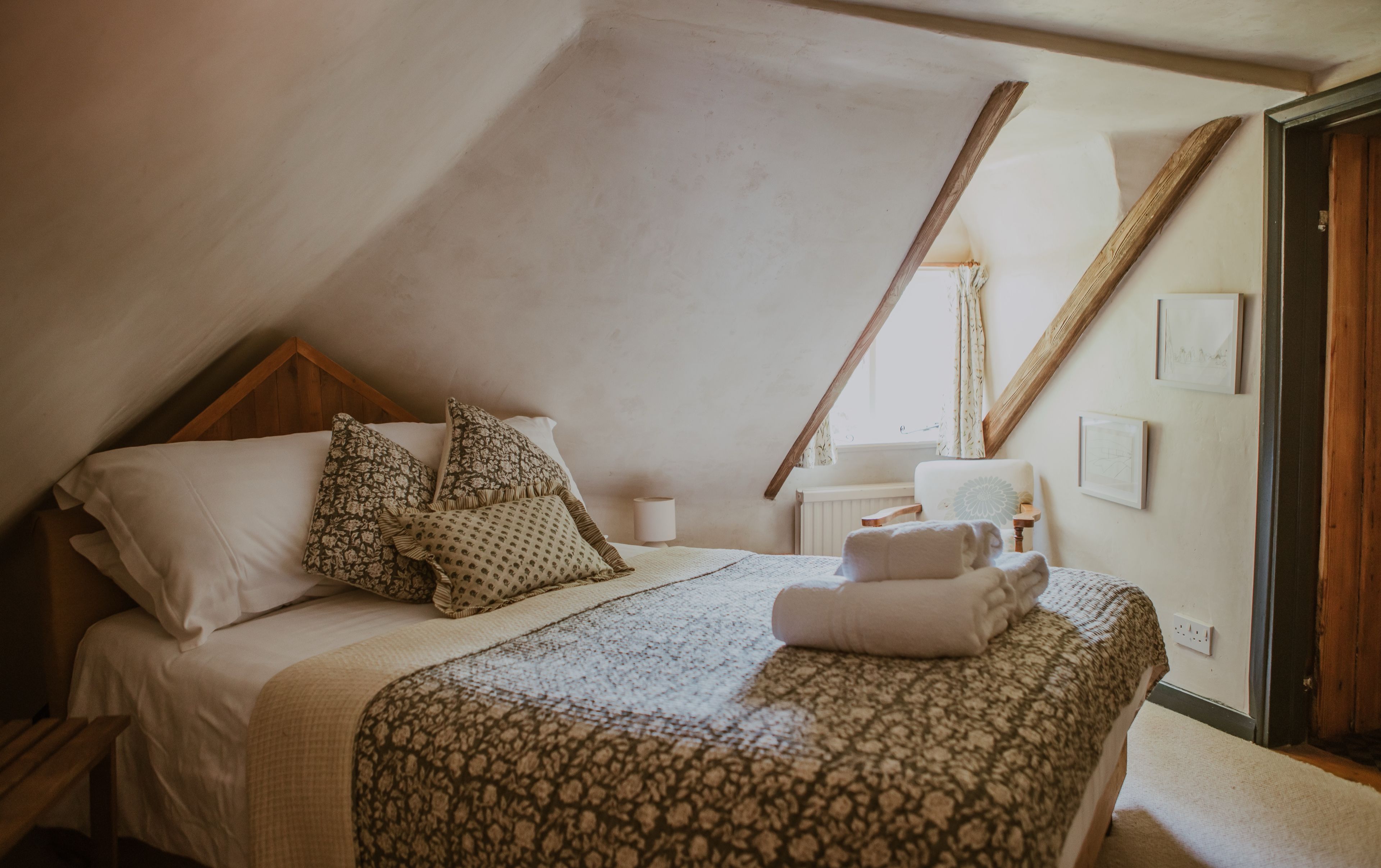 Une chambre avec un plafond en pente, un lit avec une literie à motifs, une table de chevet avec une lampe et une fenêtre avec des rideaux.