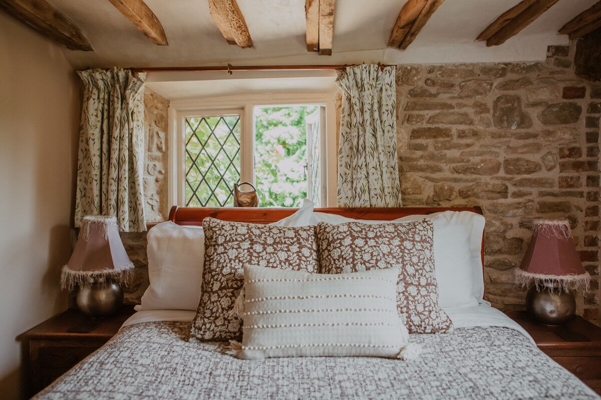 A bedroom with a stone wall, a window with curtains, a bed with patterned bedding, and two bedside lamps.