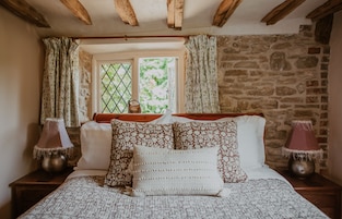 Un dormitorio con pared de piedra, una ventana con cortinas, una cama con ropa de cama estampada y dos lámparas de noche.