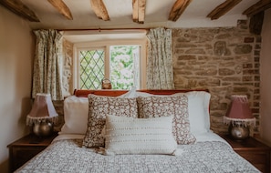 A bedroom with a stone wall, a window with curtains, a bed with patterned bedding, and two bedside lamps. - Strand House (Winchelsea)