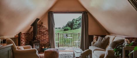 A cozy attic room with a fireplace, a sofa, a dining table, and a view of a green landscape.