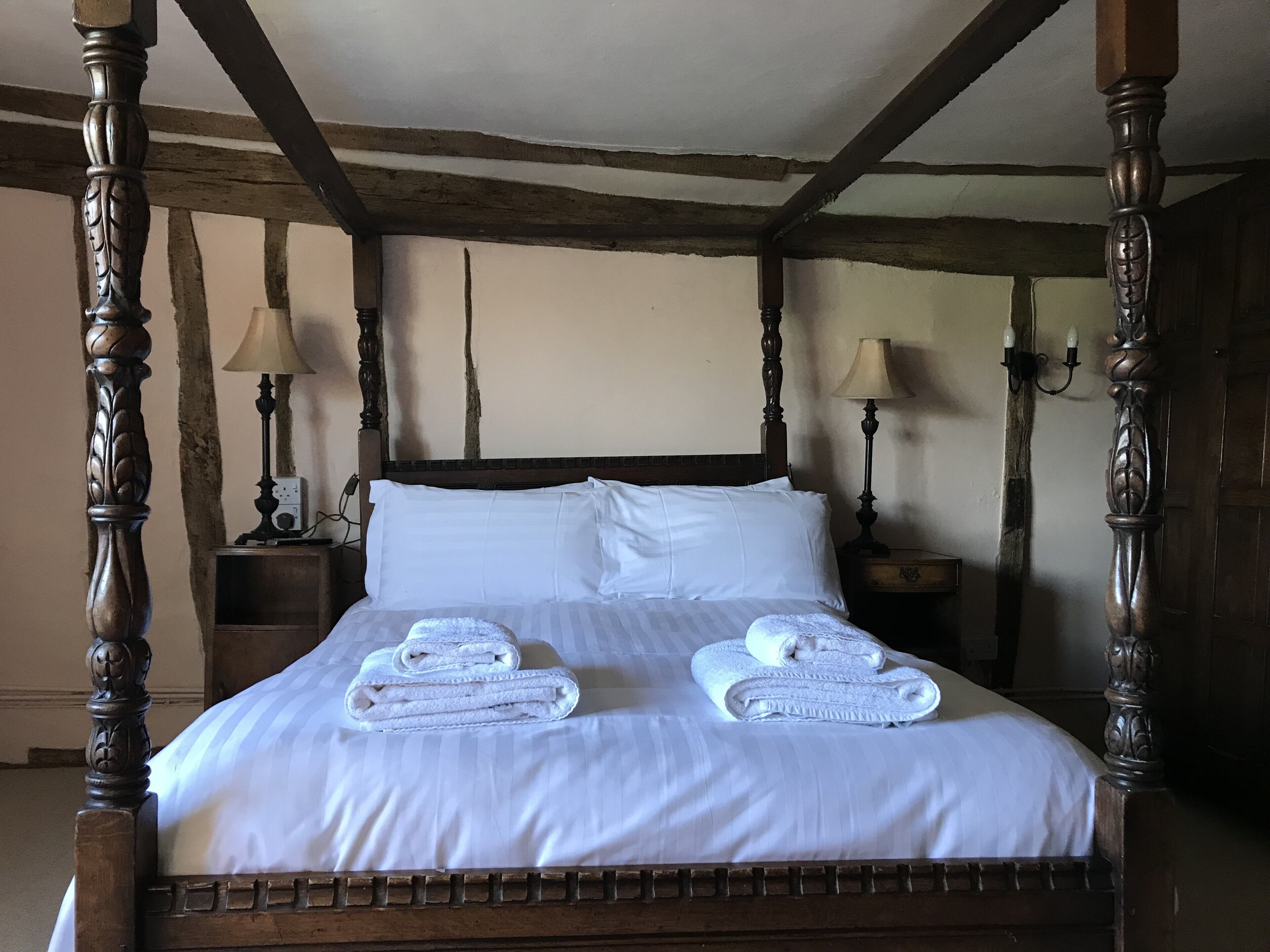 A four-poster bed with white linens and two folded towels, wooden headboard, and nightstands with lamps.