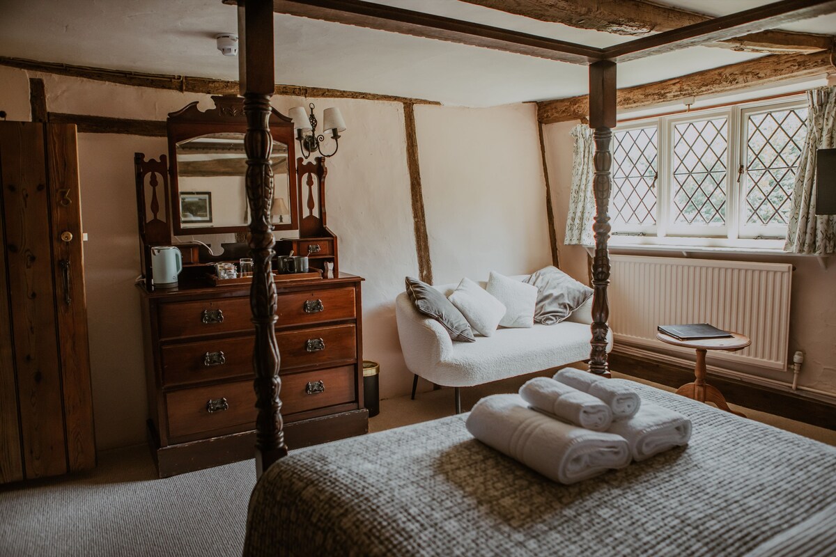 A bedroom with a four-poster bed, a wooden dresser, a mirror, and a window with a view.