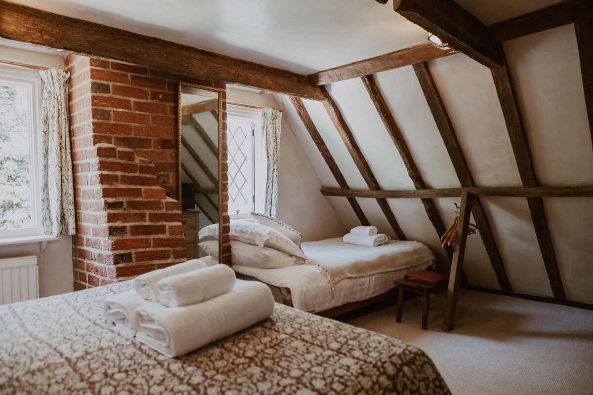 A bedroom with a sloped ceiling, exposed wooden beams, a brick wall, and a window with curtains.