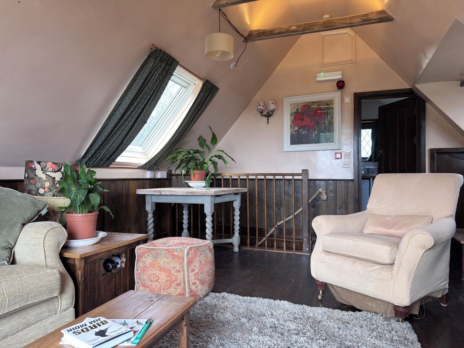 Un acogedor dormitorio tipo loft con techo inclinado, un ventanal grande con cortinas, una mesa de madera, un banco acolchado, una silla y una planta en maceta.
