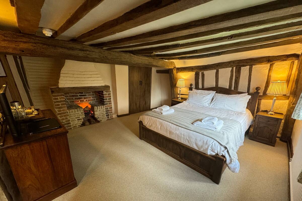 A bedroom with a large bed, a fireplace, wooden beams, a nightstand, and a lamp.