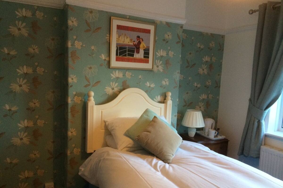 Quarto com papel de parede floral, cama branca com duas almofadas, quadro na parede e mesa de cabeceira com abajur.