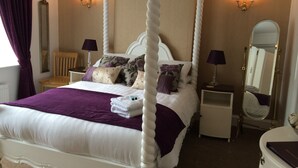 Una cama con dosel de cuatro postes con estructura blanca y ropa de cama púrpura, dos mesitas de noche con lámparas, un espejo y una silla de madera.