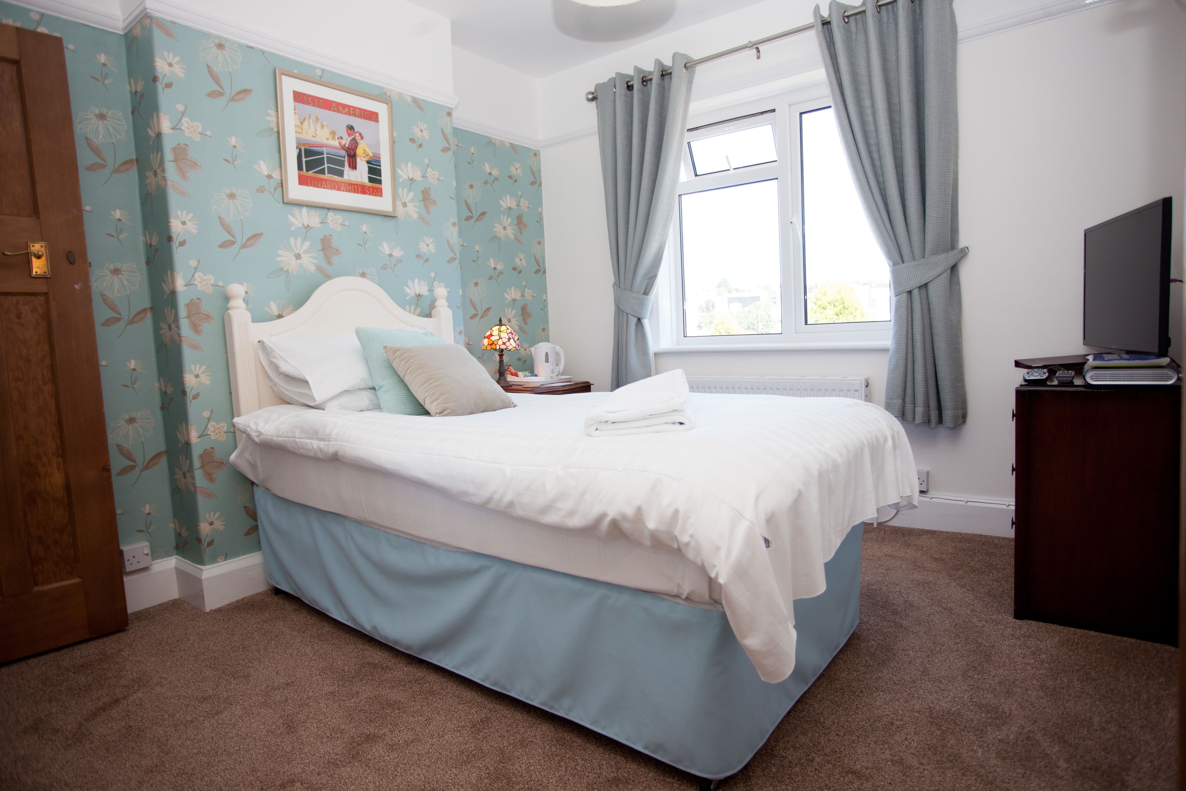 A bedroom with floral wallpaper, a bed with white bedding, a nightstand with a lamp, and a window with curtains.
