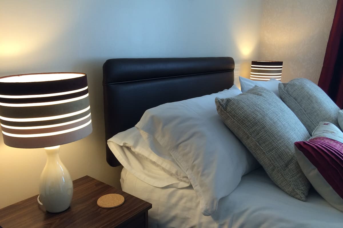 Cama bem arrumada com lençóis brancos e várias almofadas, cabeceira escura e duas luminárias de cabeceira com cúpulas listradas.