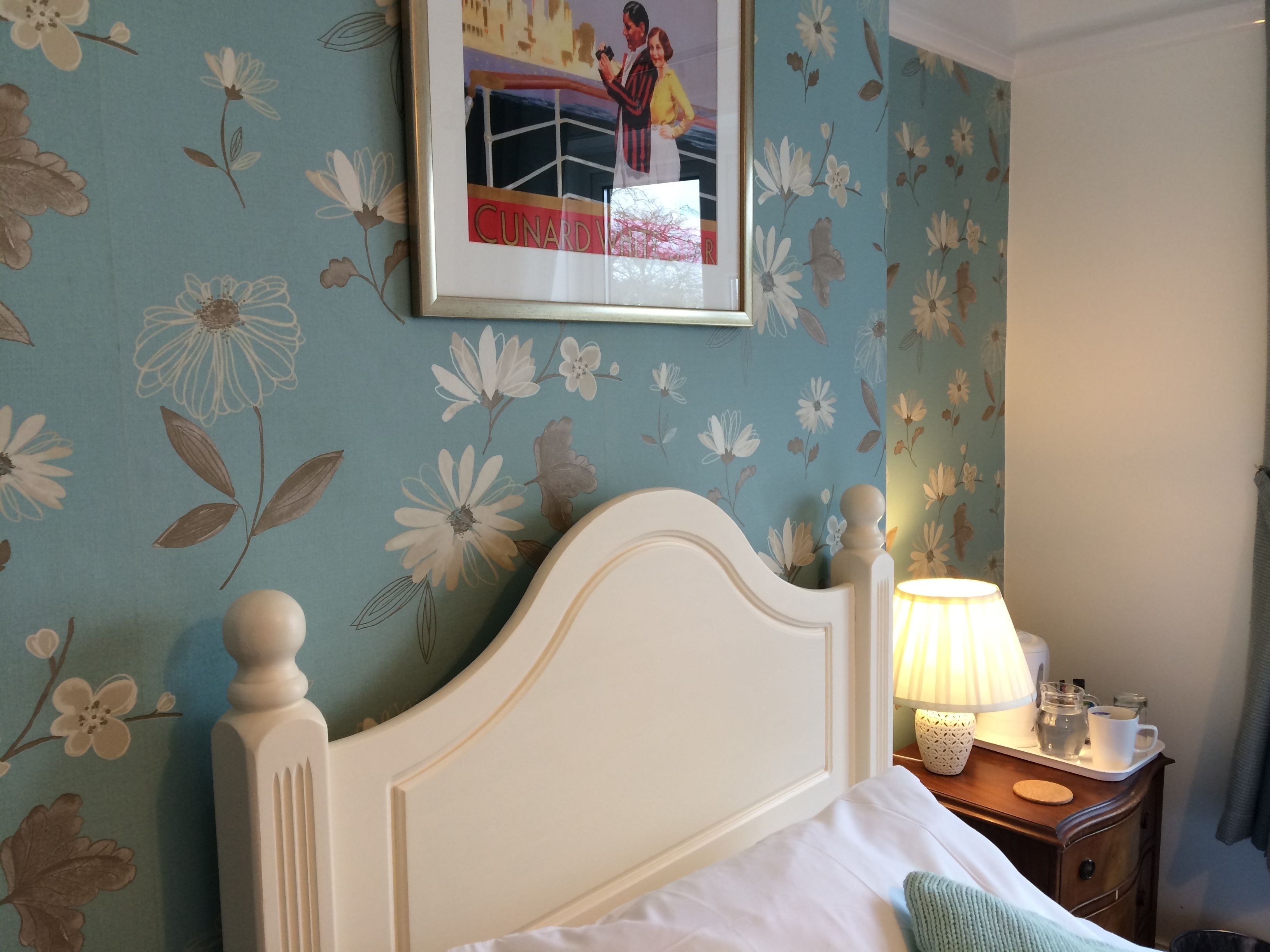 Une chambre à coucher avec un papier peint à motifs floraux, un lit blanc avec une tête de lit, une table de chevet avec une lampe et un tableau encadré accroché au mur.