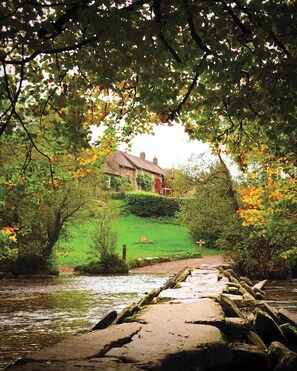 A stone bridge over a river leading to a house surrounded by greenery.