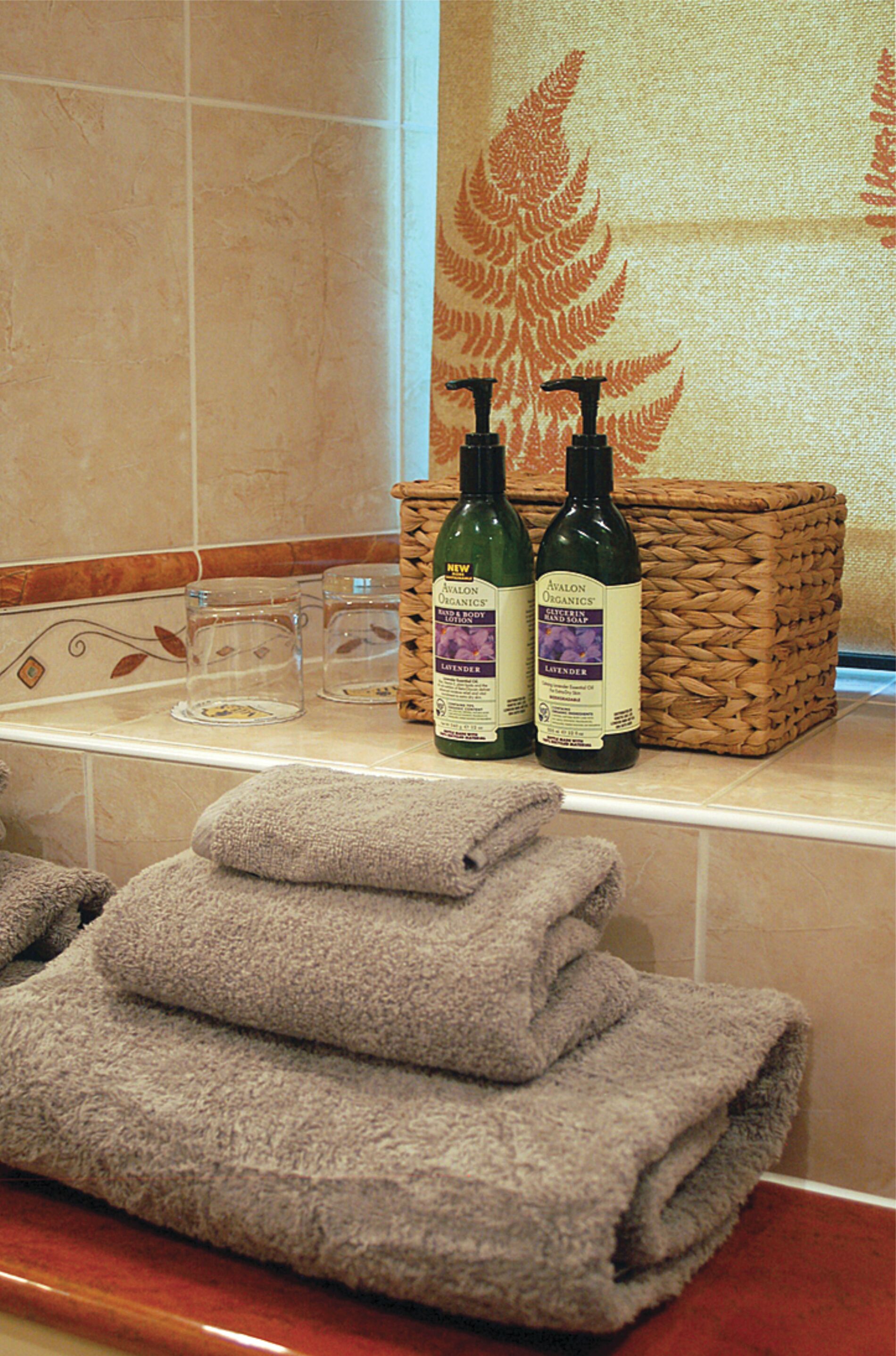Un bagno con parete piastrellata, un cesto in vimini, due flaconi di lozione corpo alla lavanda Avalon Organics e tre asciugamani piegati.
