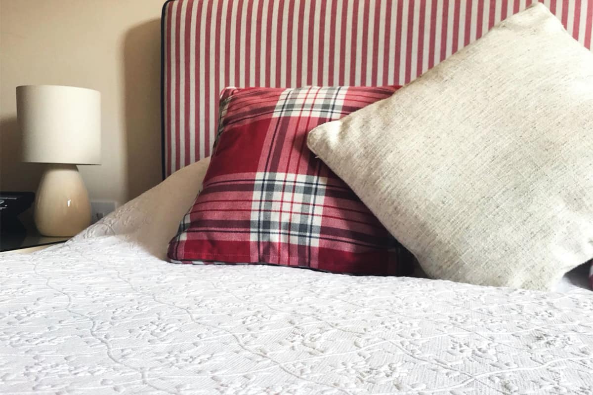 A bed with a white quilt, a red and black plaid pillow, and a beige pillow.