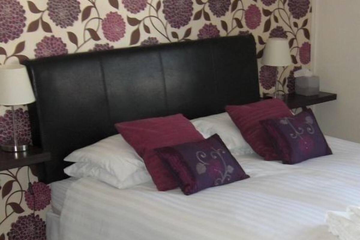A neatly made bed with white linens and two purple pillows, a dark headboard, and a wall with a floral pattern.
