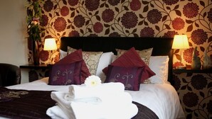 A neatly made bed with a dark headboard, two lamps on either side, and a floral wallpaper pattern. - The Bell (Pershore)