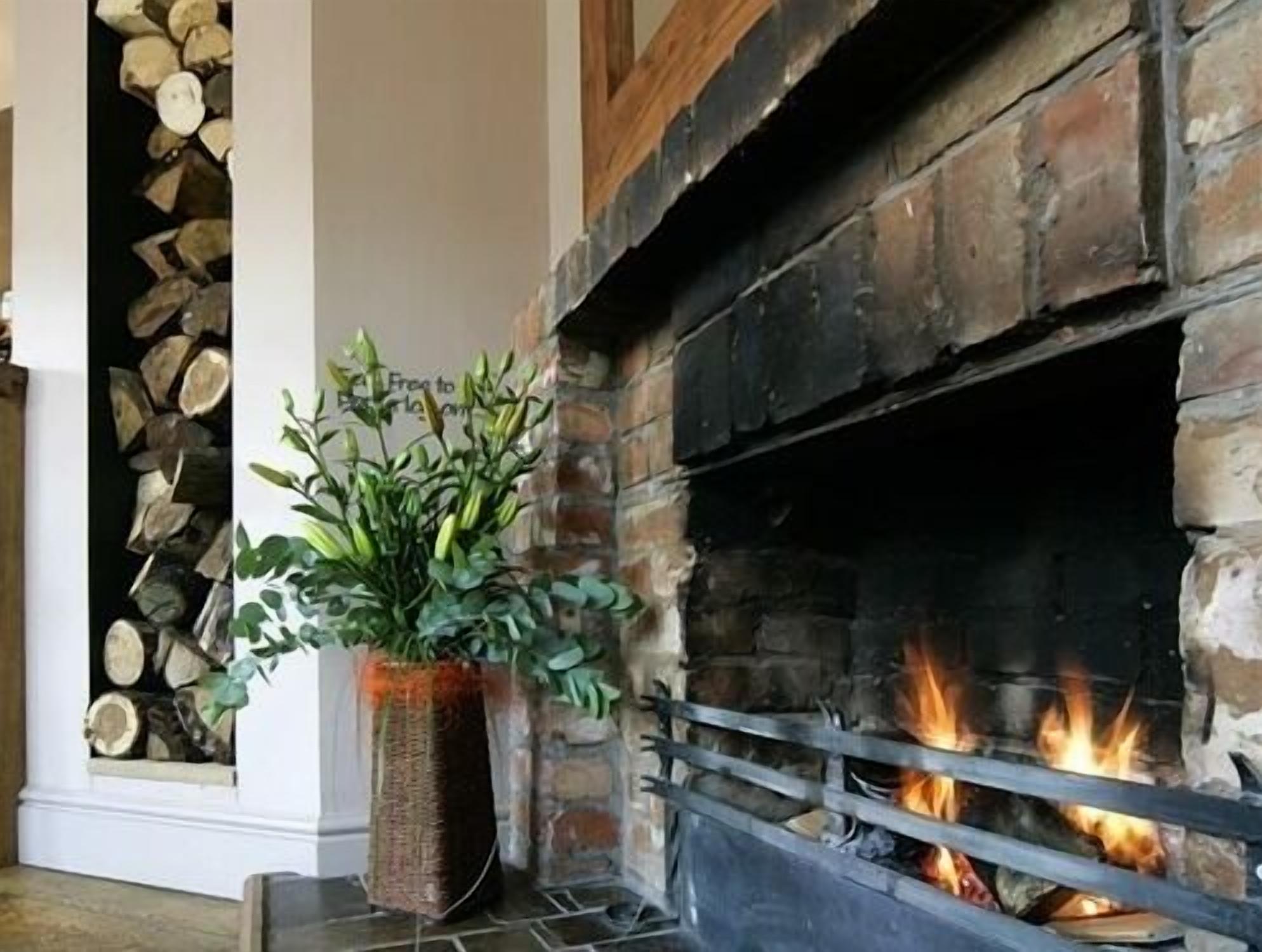 A fireplace with a fire burning, surrounded by a stone wall and a wooden mantel.