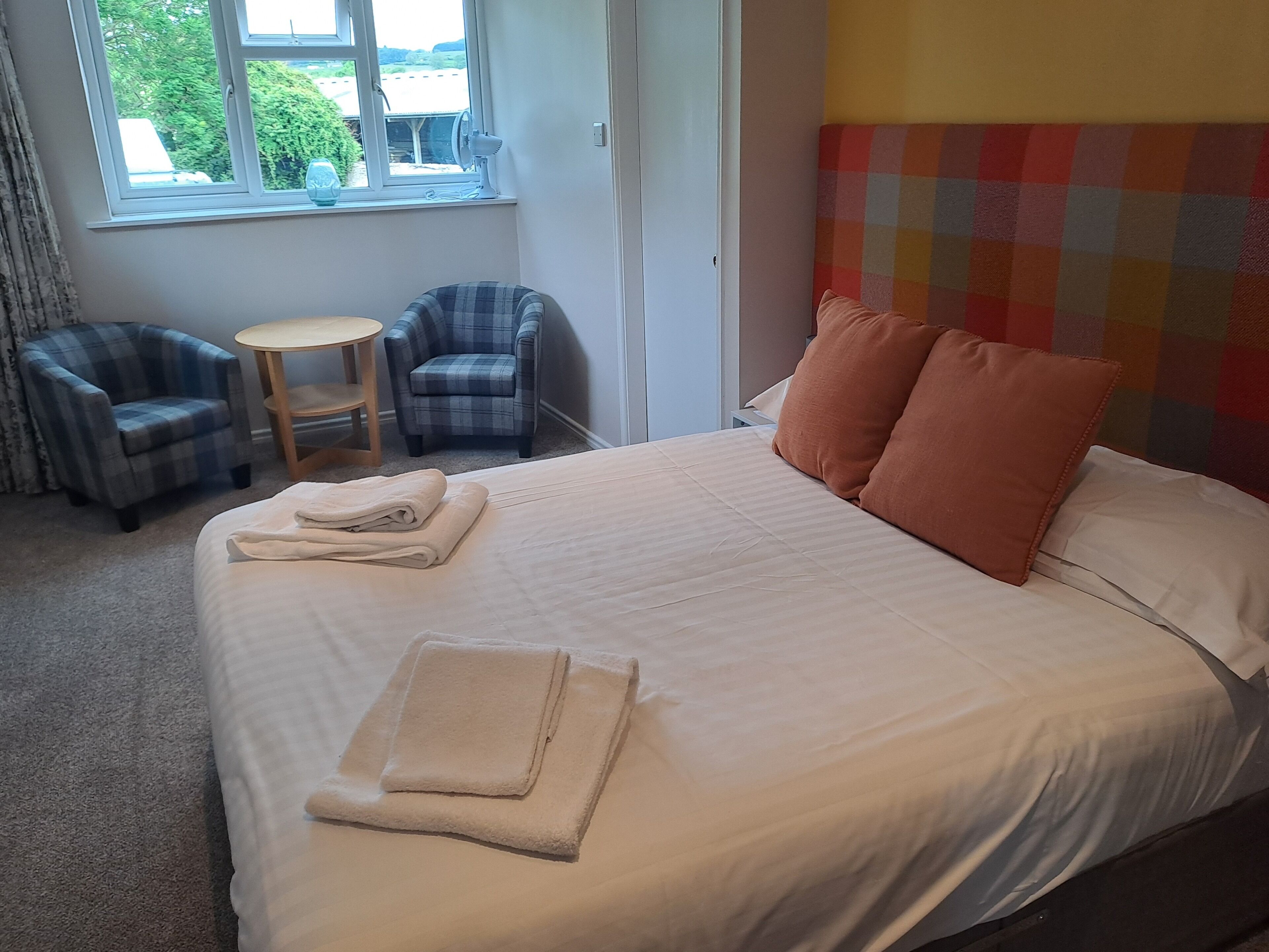Een hotelkamer met een bed, twee stoelen, een klein tafeltje en een raam met uitzicht op groen.