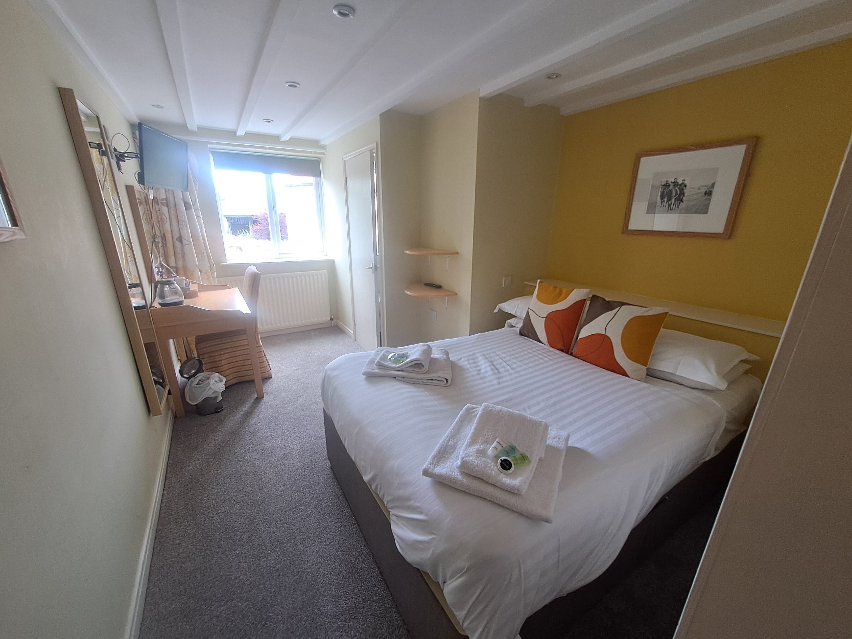 Habitación de hotel con cama, escritorio, silla, radiador, ventana y un cuadro en la pared.