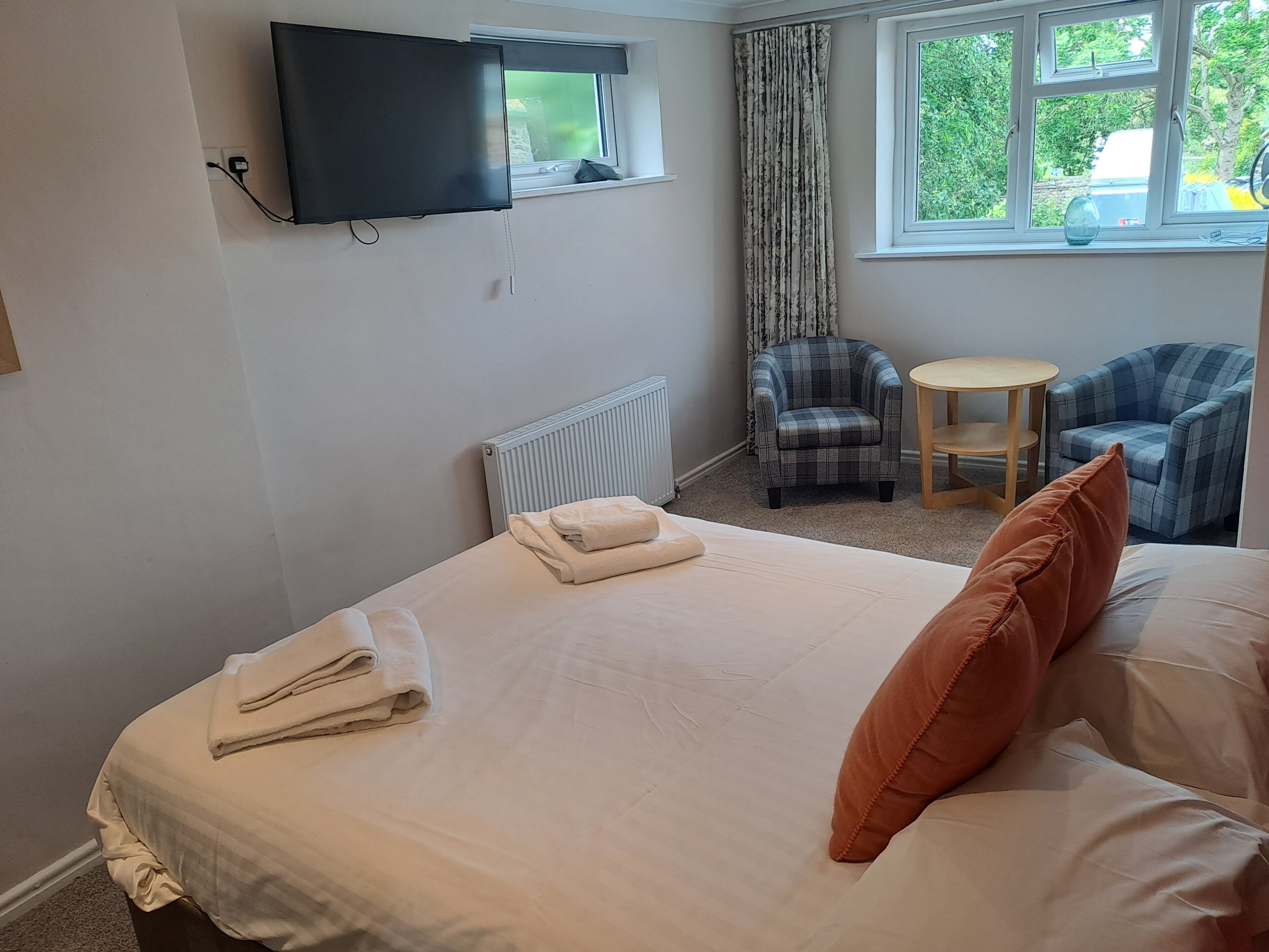 Een hotelkamer met een bed, een flatscreen televisie aan de muur, een raam met gordijnen, een radiator, twee geruite fauteuils en een klein rond tafeltje.