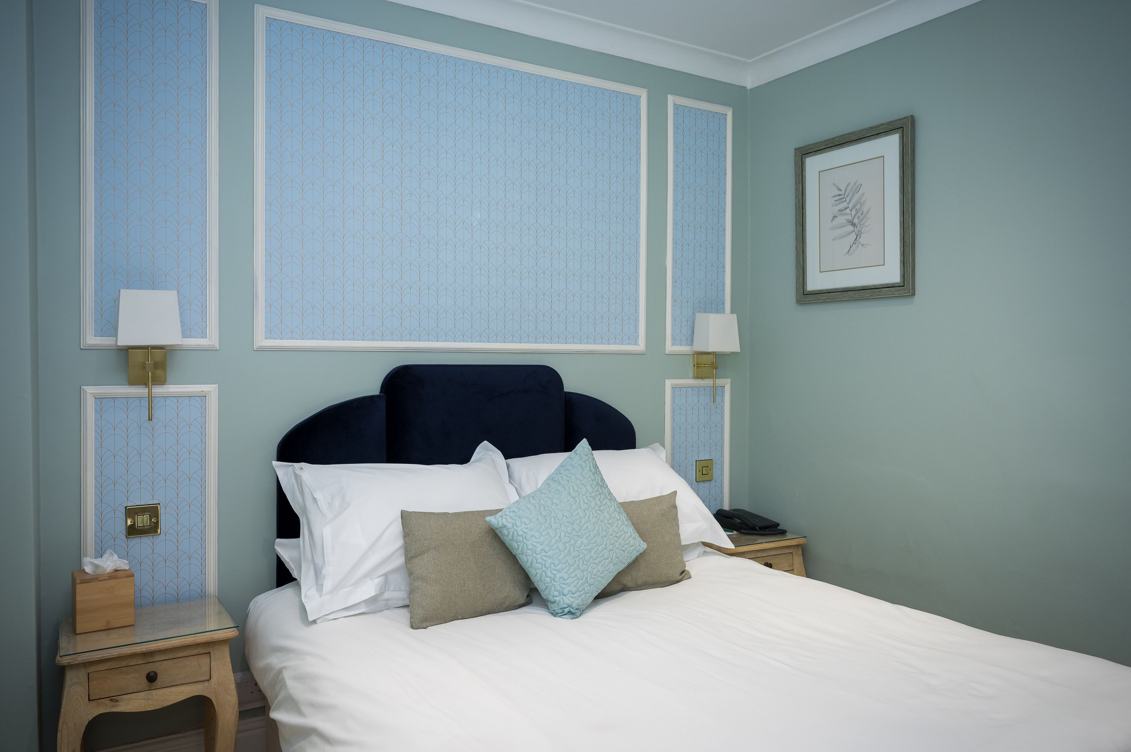 A neatly made bed with white and beige pillows, a dark headboard, and a bedside table with a lamp and a tissue box.