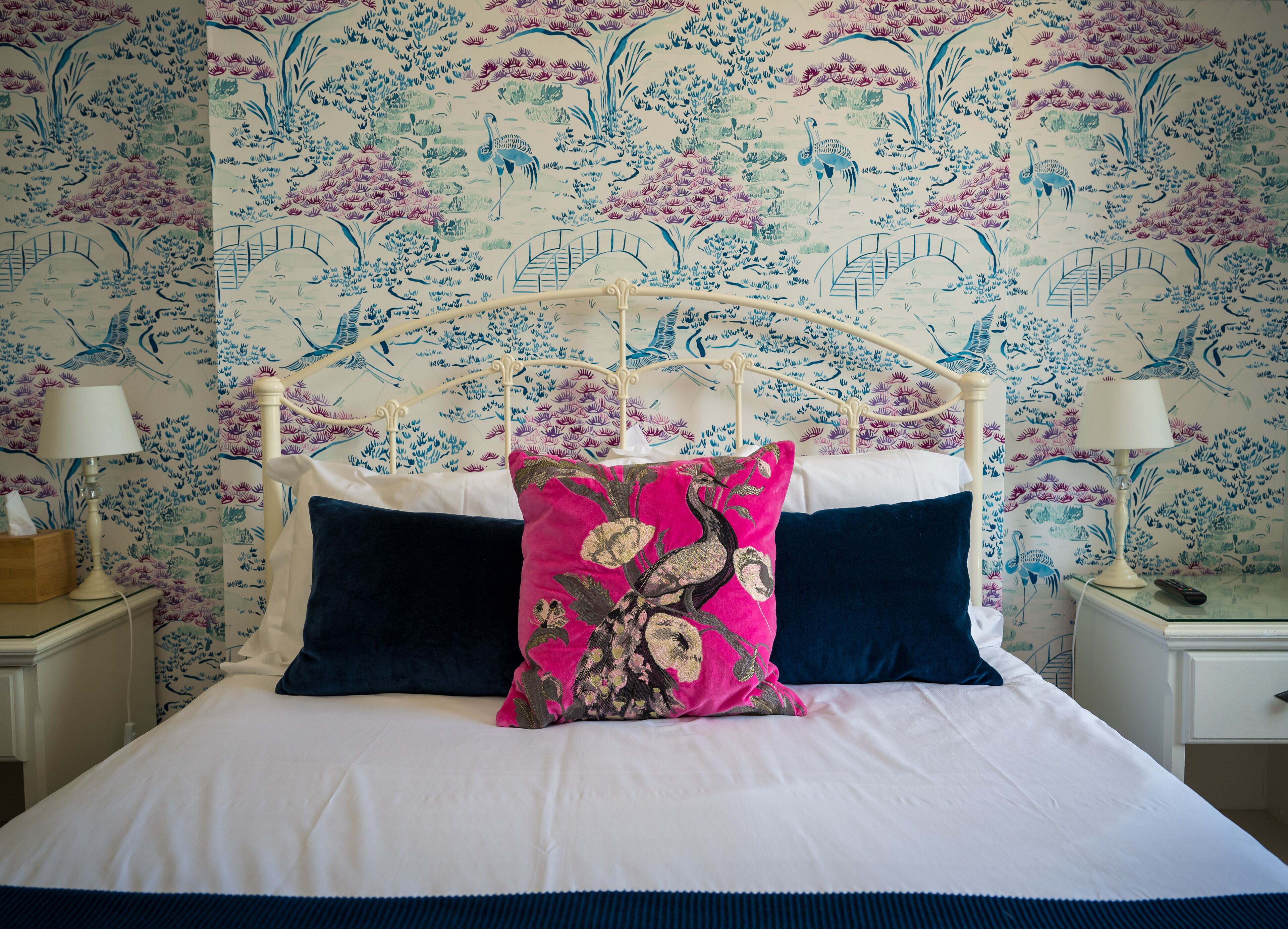 Una cama con colcha blanca y azul marino, un cojín floral rosa y cojines azul marino. Hay dos mesitas de noche con lámparas y una mesita de noche con una lámpara. El papel tapiz de la pared detrás de la cama presenta un estampado floral.