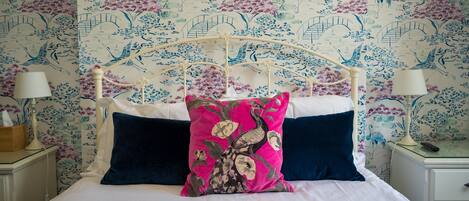 Una cama con colcha blanca y azul marino, un cojín floral rosa y cojines azul marino. Hay dos mesitas de noche con lámparas y una mesita de noche con una lámpara. El papel tapiz de la pared detrás de la cama presenta un estampado floral.