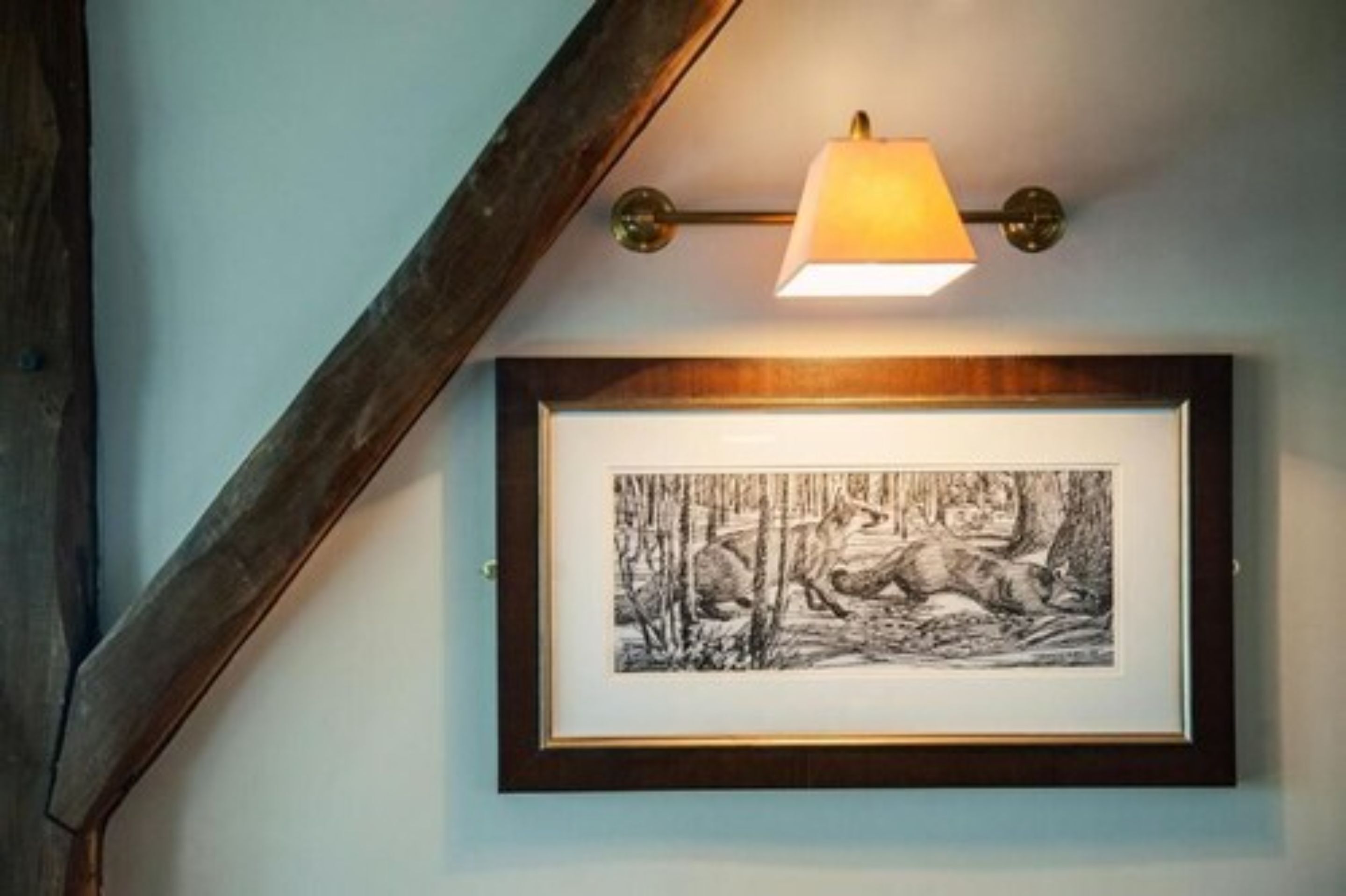 A framed black and white drawing of a wolf and a deer on a wall, with a wall-mounted lamp above it.