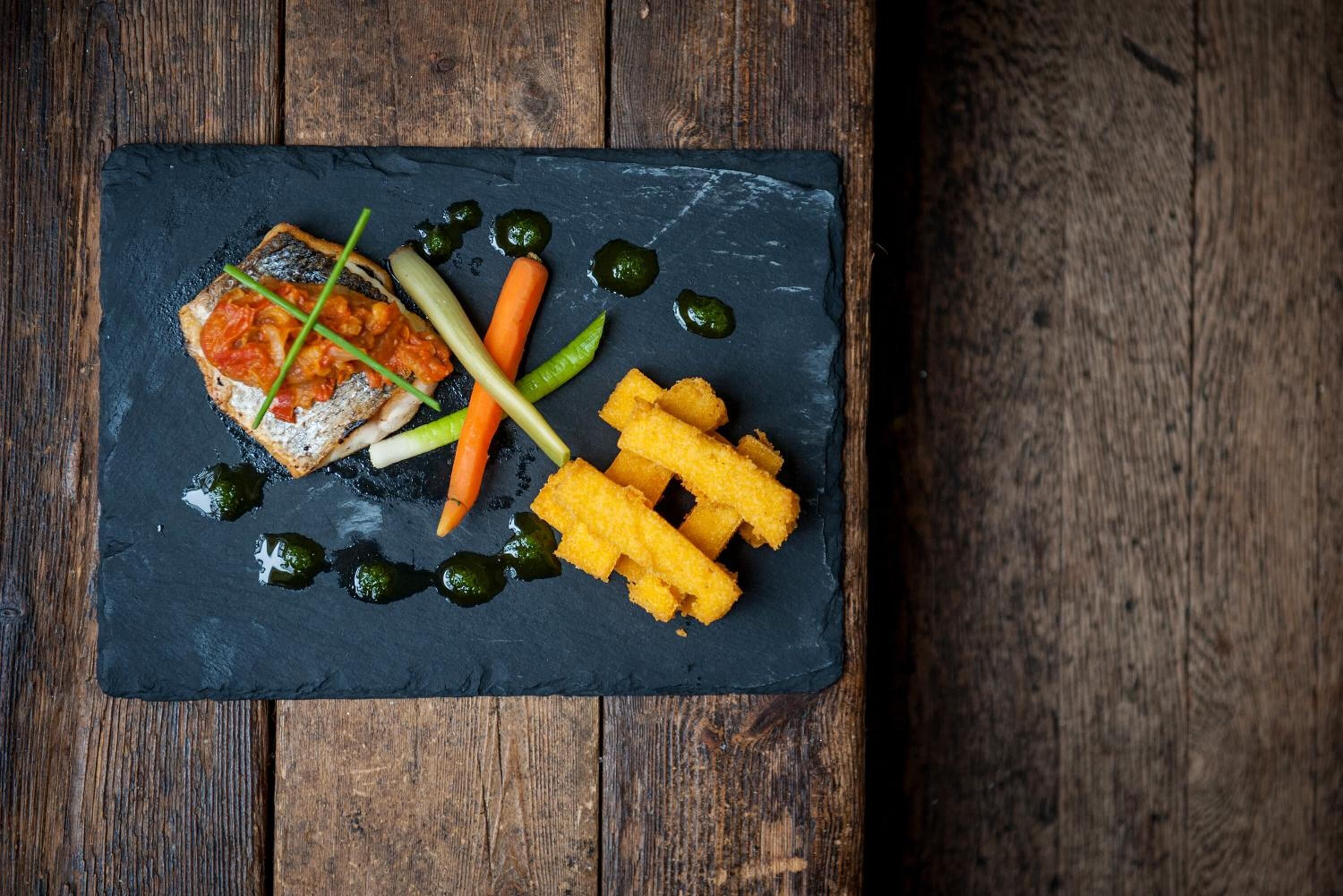 Une planche de service en ardoise noire, sur laquelle on a disposé un morceau de poisson, des olives vertes, des carottes marinées et des haricots verts marinés, le tout posé sur une table en bois.