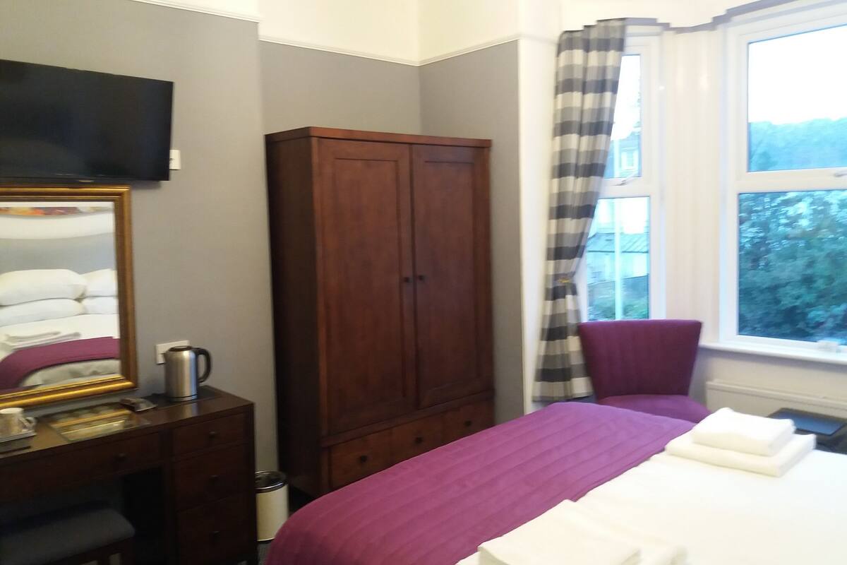 A hotel room with a bed, a wooden wardrobe, a desk with a mirror, a television, and a window with curtains.