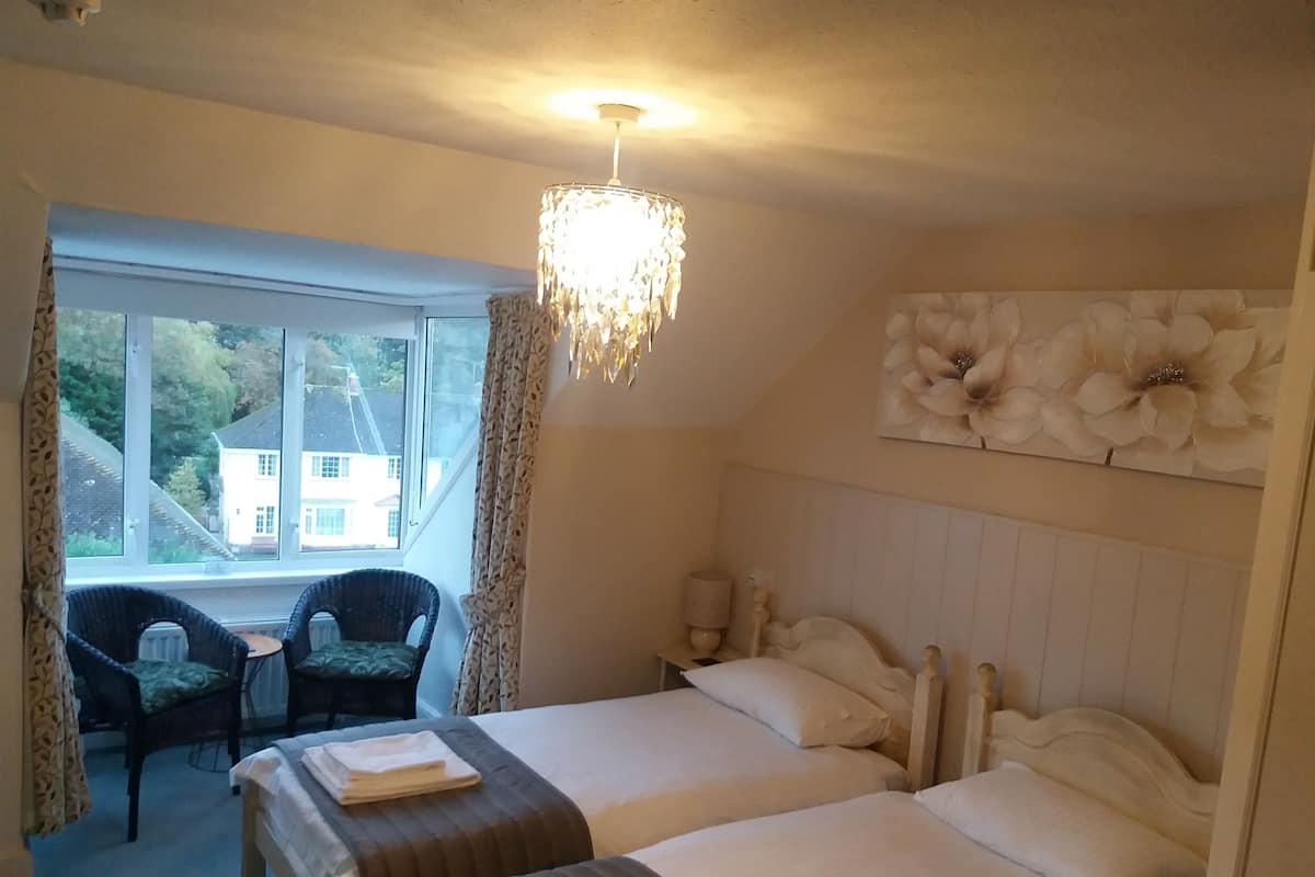 A hotel room with two beds, a chandelier, a view of houses outside the window, and a chair by the window.